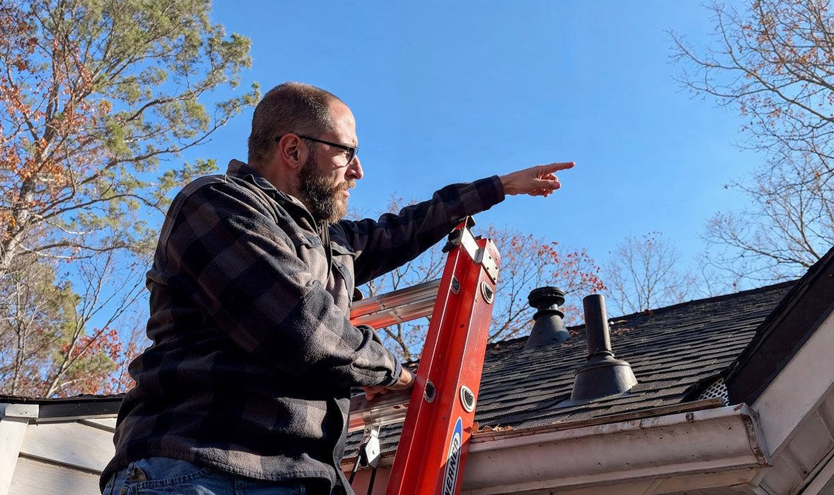 Man in plaid jacket pointing at something while standing on a red ladder next to a house roof with chimney, clear blue sky in the background.
